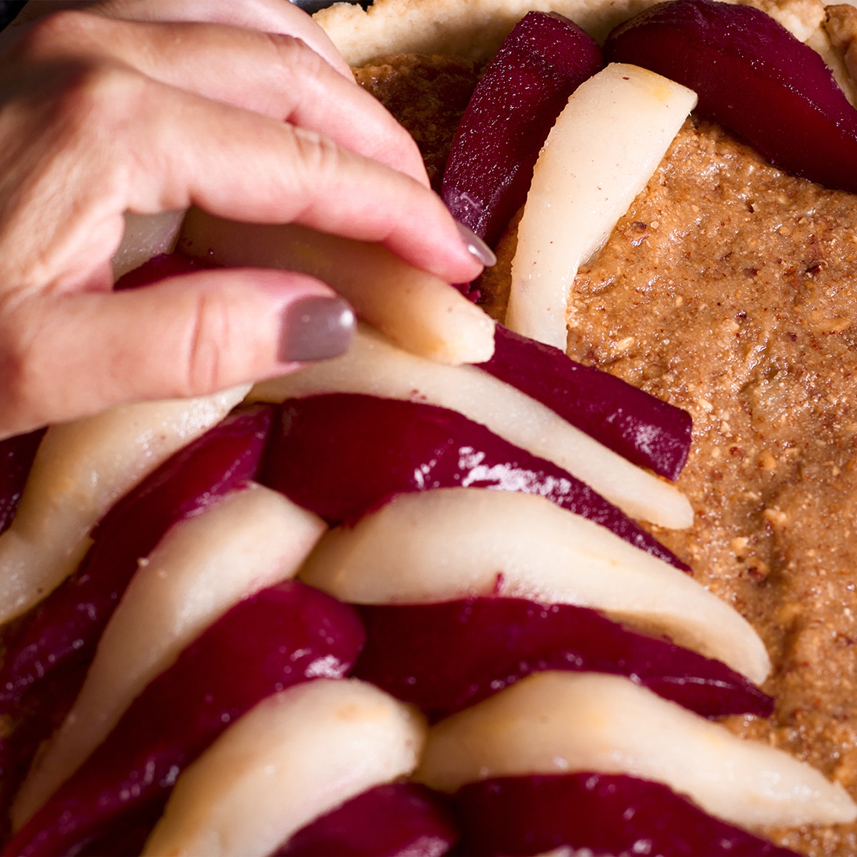 Someone using their hand to arrange wine poached pears over a tart filled with frangipane. 