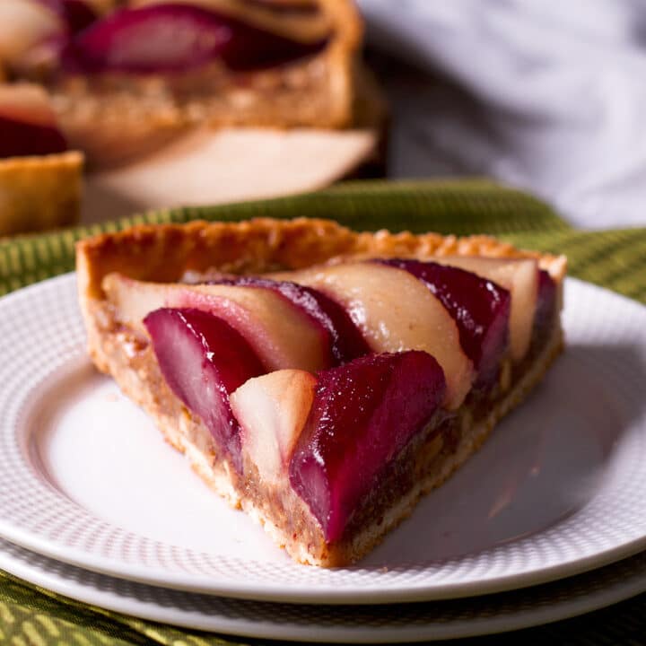 A slice of wine poached pear frangipane tart on a white plate with the rest of the tart in the background.