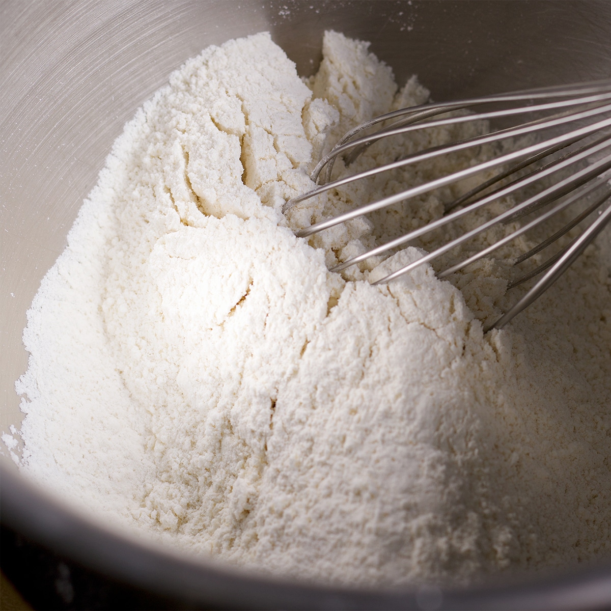 Someone using a wire whisk to mix flour and sugar in a mixing bowl.