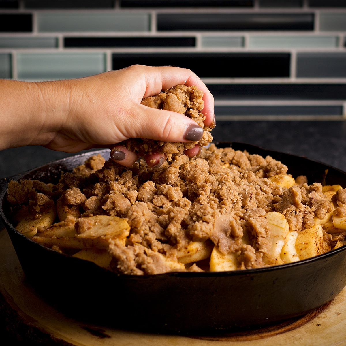 Someone using their hands to top apple cobbler filling in a cast iron skillet with brown sugar cookie topping.
