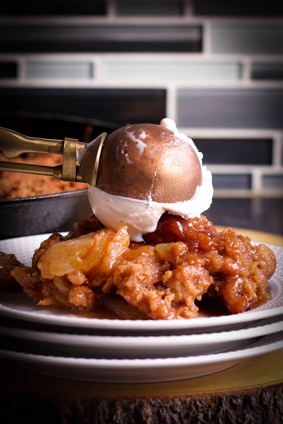 Someone using an old fashioned ice cream scoop to top a serving of apple cobbler with a scoop of vanilla ice cream.