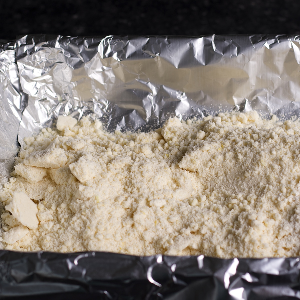 Adding the crumbly raw shortbread dough to a pan that's been lined with aluminum foil.