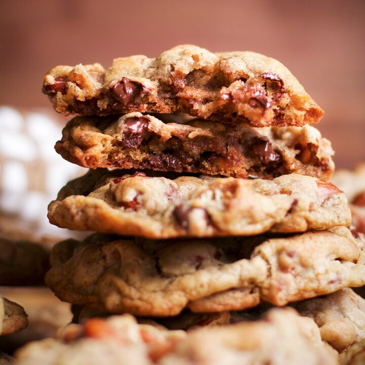 A stack of several cookies on a table with the top cookie broken open to reveal chocolate chips and nuts.