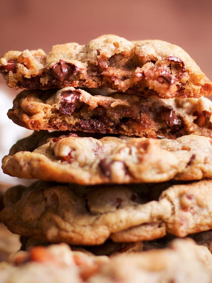 A stack of several cookies on a table with the top cookie broken open to reveal chocolate chips and nuts.