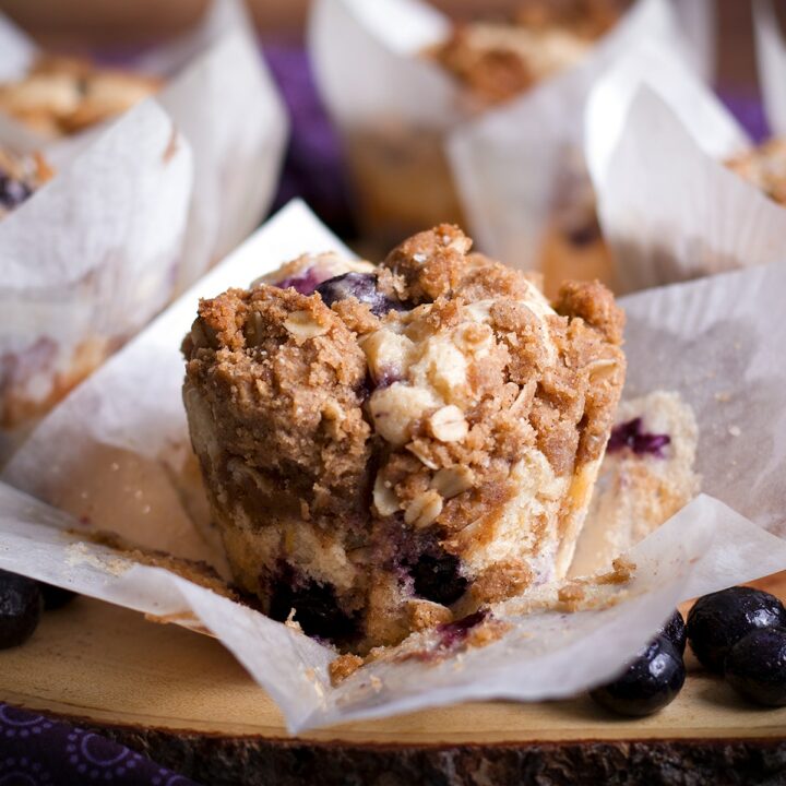 A sour cream streusel blueberry muffin on a wooden tray surrounded by more muffins.