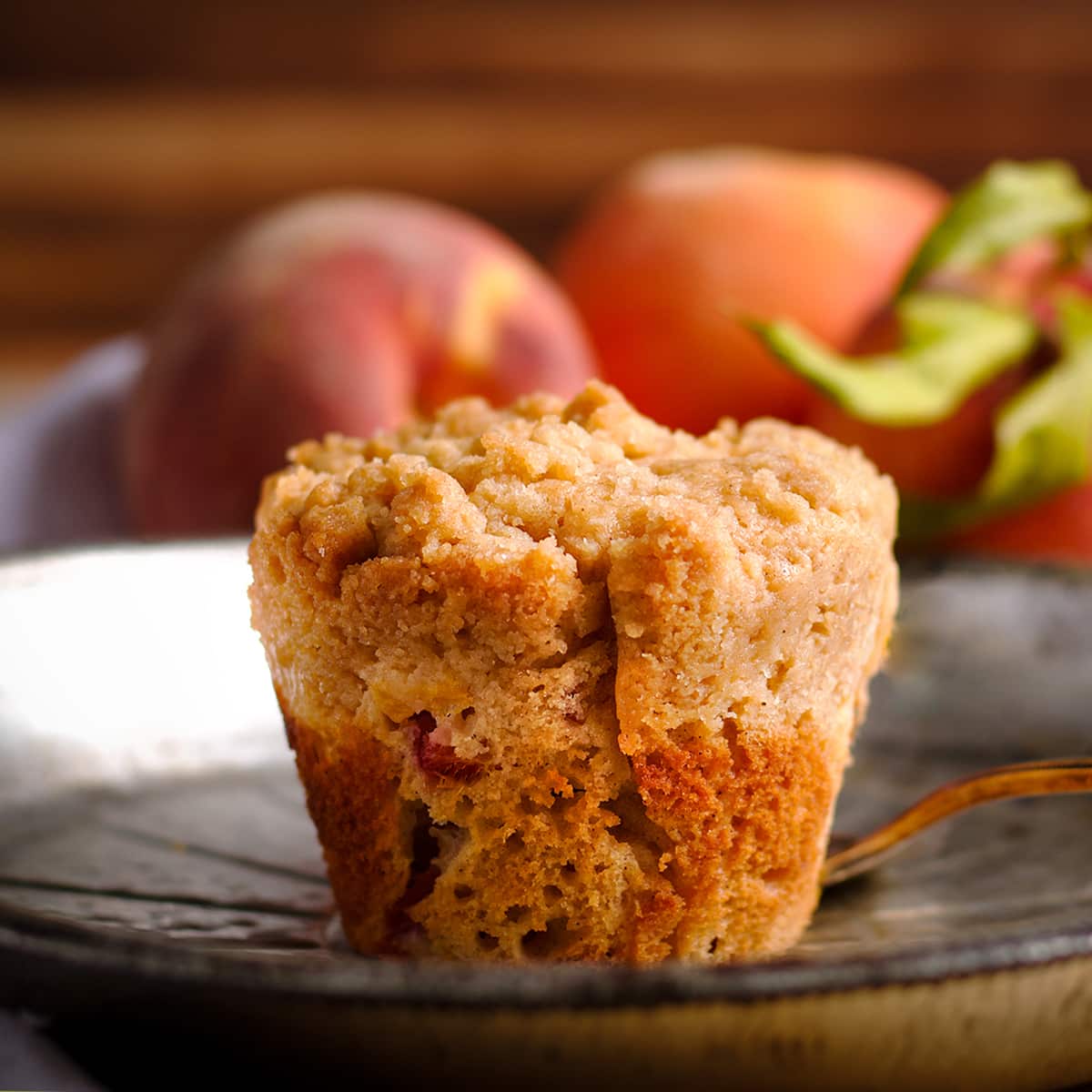 A peach cobbler muffin sitting on a tin plate with fresh peaches in the background.