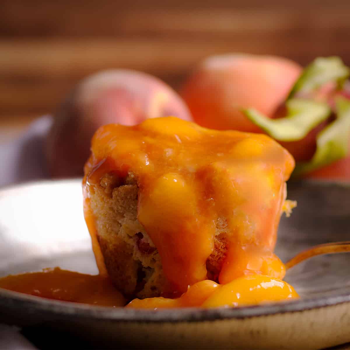 A peach cobbler muffin smothered with peach sauce on a tin plate with fresh peaches in the background.