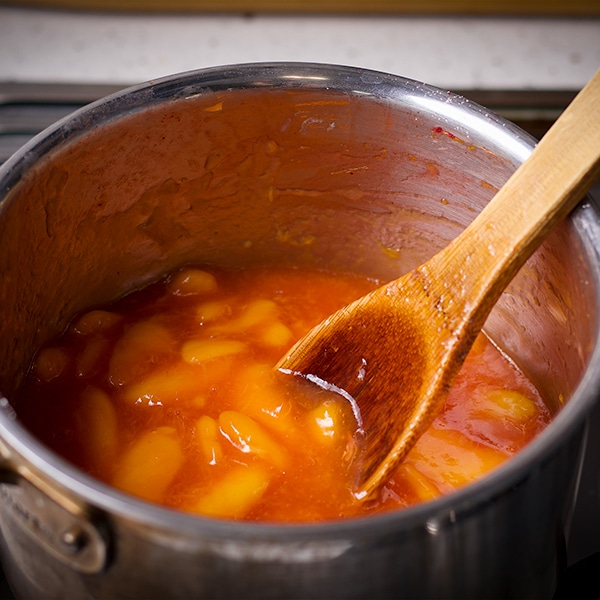 Someone using a wood spoon to stir a saucepan filled with peach sauce while it cooks on the stovetop.