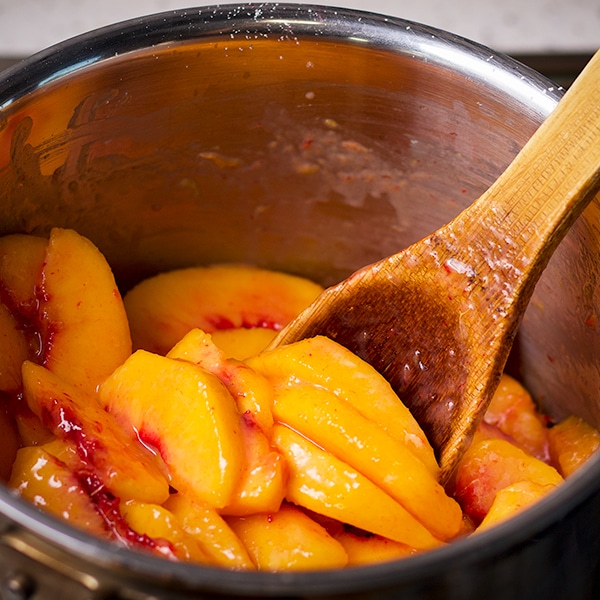 Someone using a wood spoon to stir sliced peaches in a saucepan.