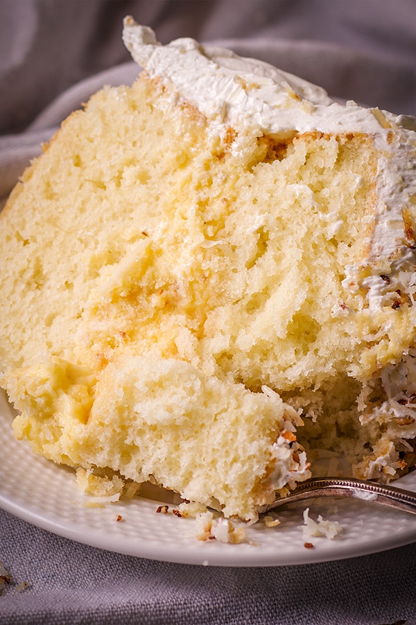 A thick slice of coconut cream cake on a white plate with a gold fork cutting a bite from the cake.