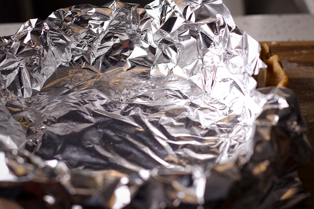A pie plate lined with toasted almond pie crust dough that's been covered in a sheet of aluminum foil.