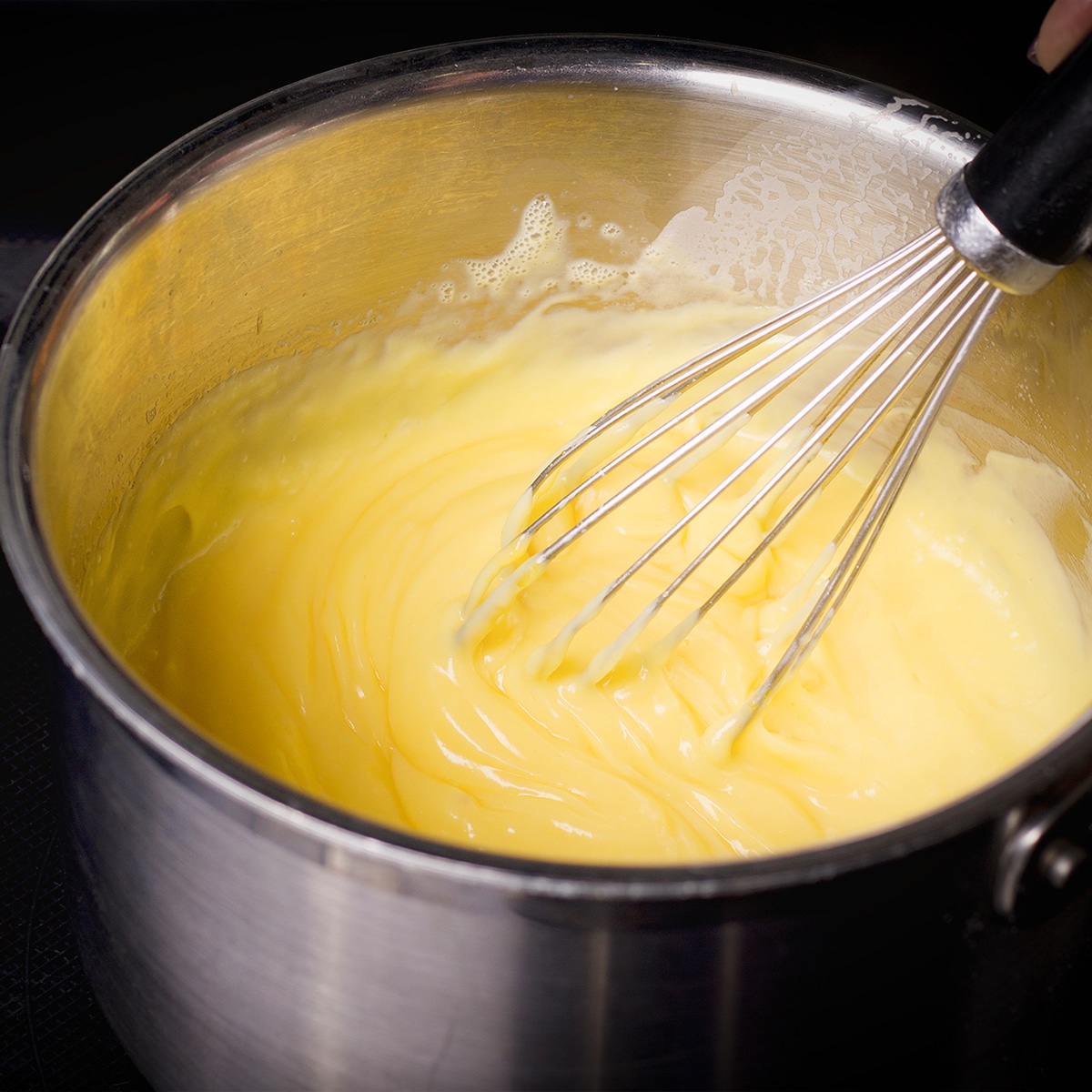 Using a whisk to stir pastry cream as it cooks and thickens in a saucepan.