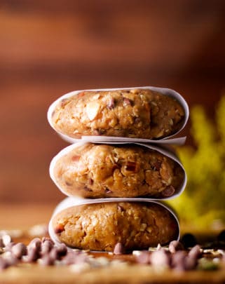 Three parchment wrapped no-bake, high protein chocolate peanut butter breakfast cookies stacked on top of each other on a wooden tray.