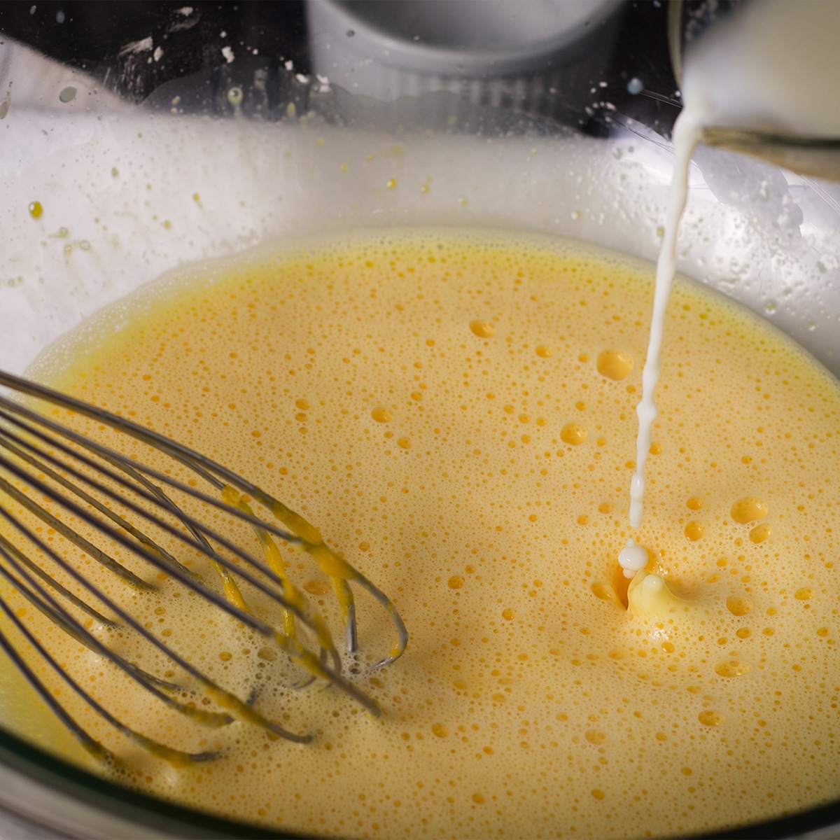 Pouring hot milk into egg yolks while stirring constantly with a wire whisk.