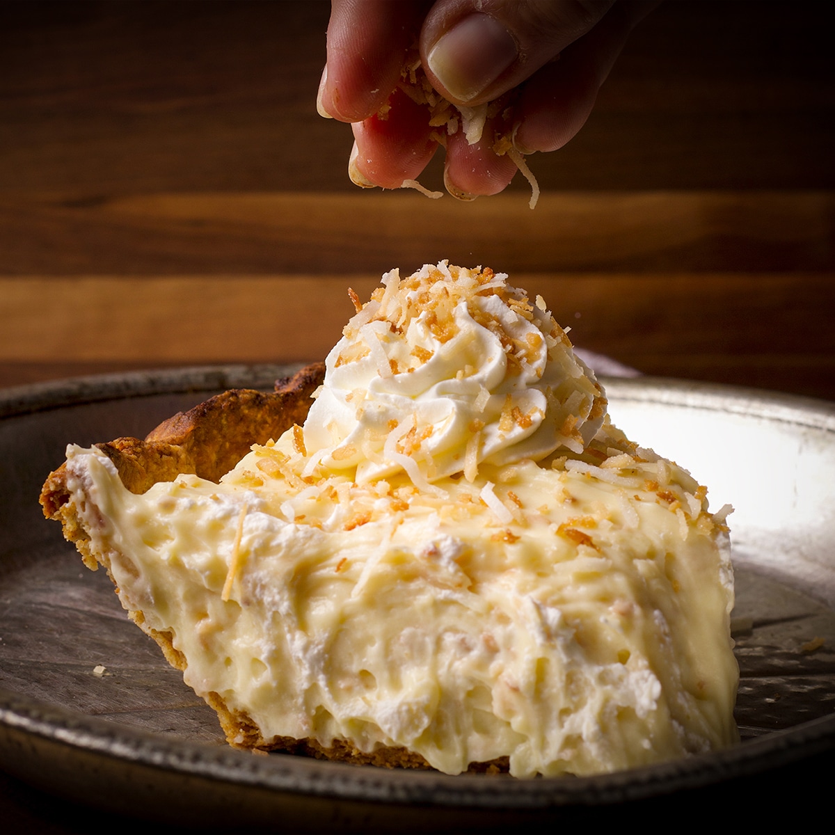 Someone sprinkling toasted coconut over a slice of coconut cream pie.
