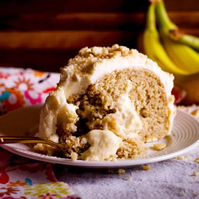 A slice of banana cream cake on a white plate with a bunch of bananas in the background. A gold fork is cutting a bite from the slice of cake.