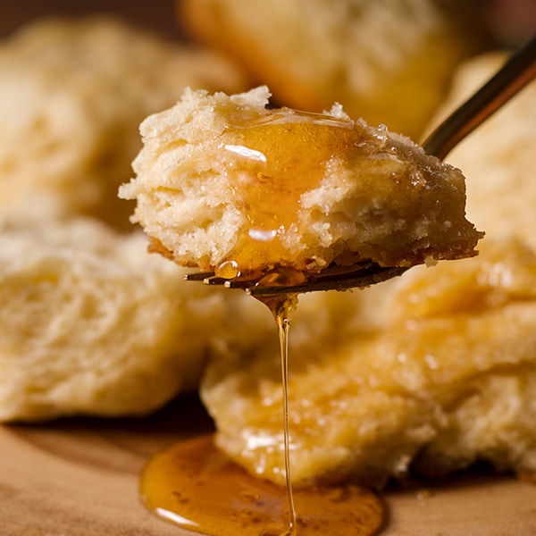 A fork with a bite of buttermilk biscuit on it that's dripping with honey.