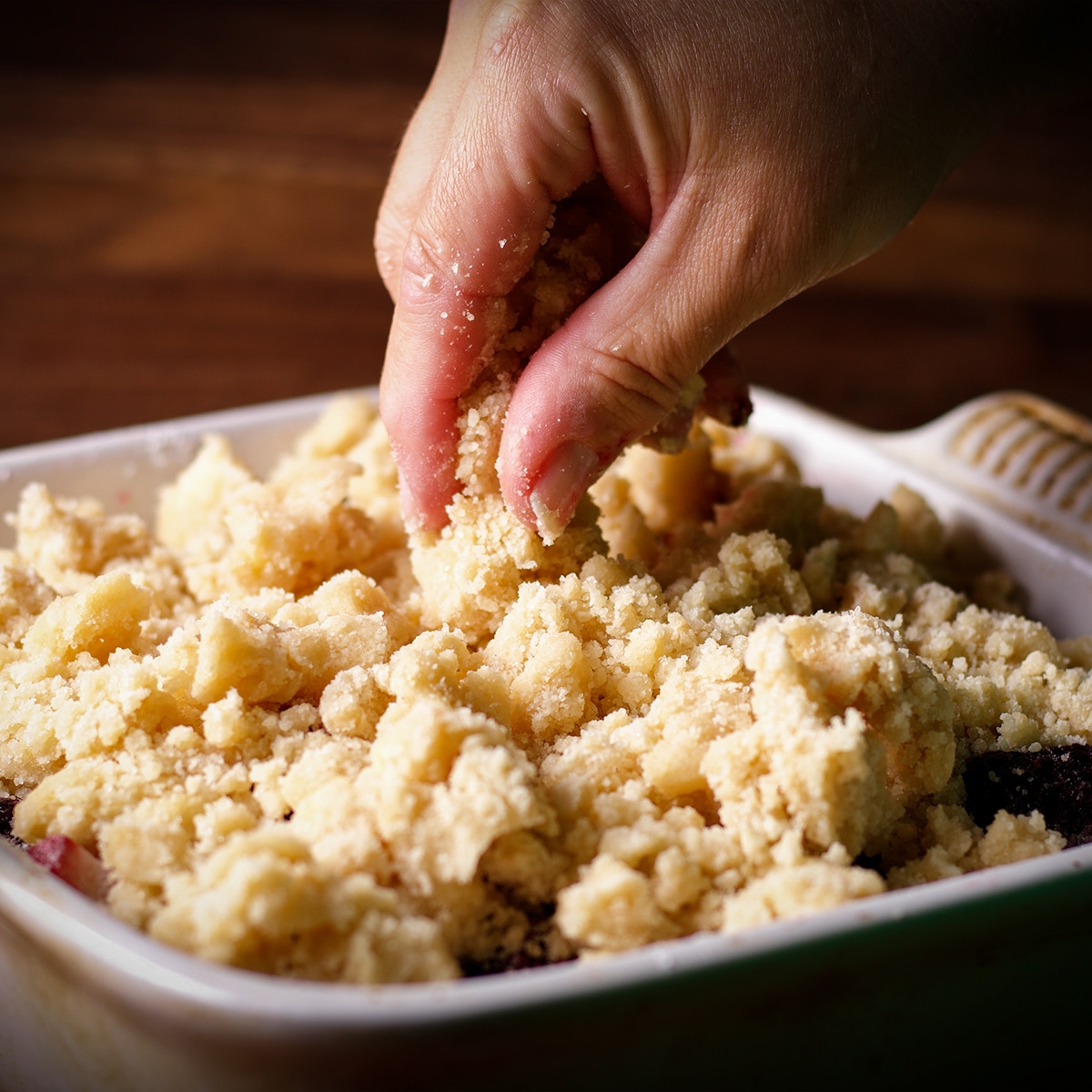 Someone using their fingers to distribute the topping for berry cobbler over the mixed berry filling.