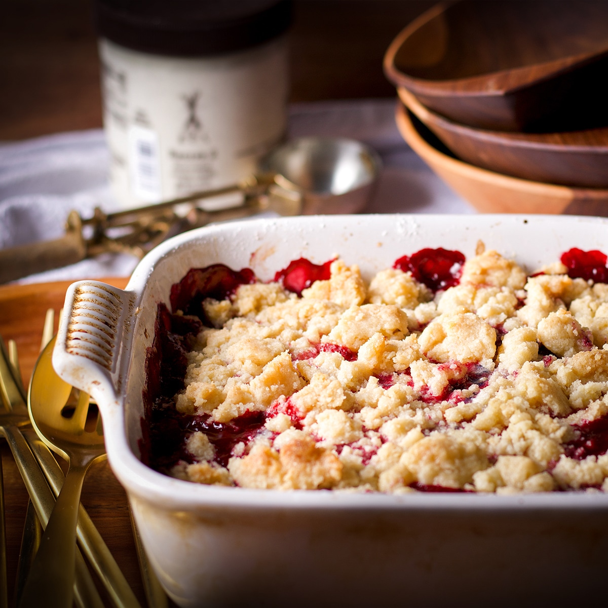 A pan of freshly baked berry cobbler cooling on a wood cutting board with bowls, spoons, and a container of vanilla ice cream near by.