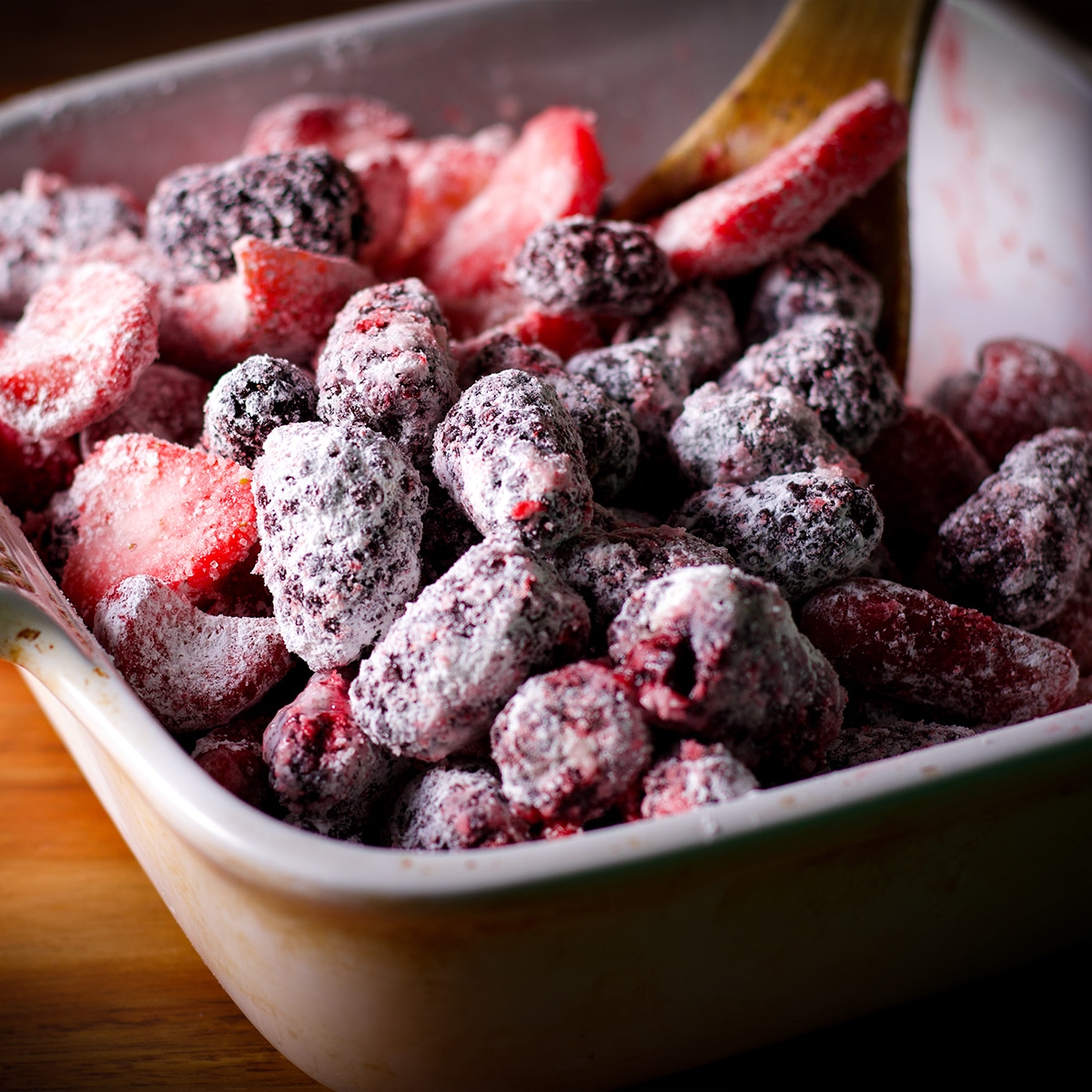 Using a wooden spoon to mix the berry topping for berry cobbler in a white baking dish.