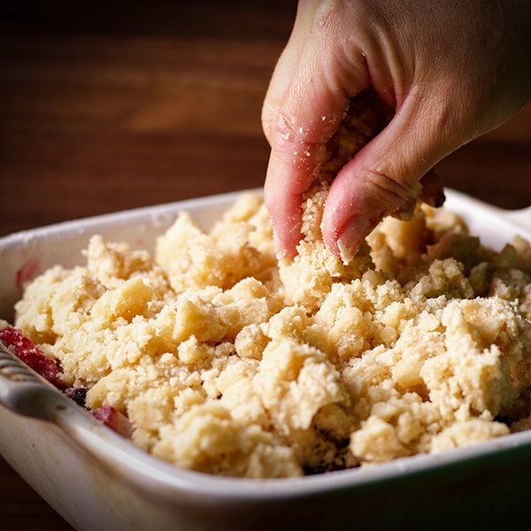 Someone using their fingers to distribute the topping for berry cobbler over the filling.