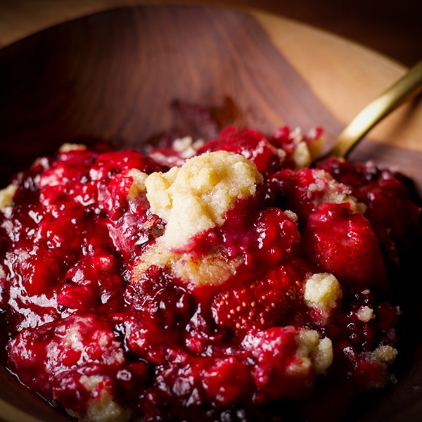 A wood bowl filled with berry cobbler and a gold spoon, ready to eat.