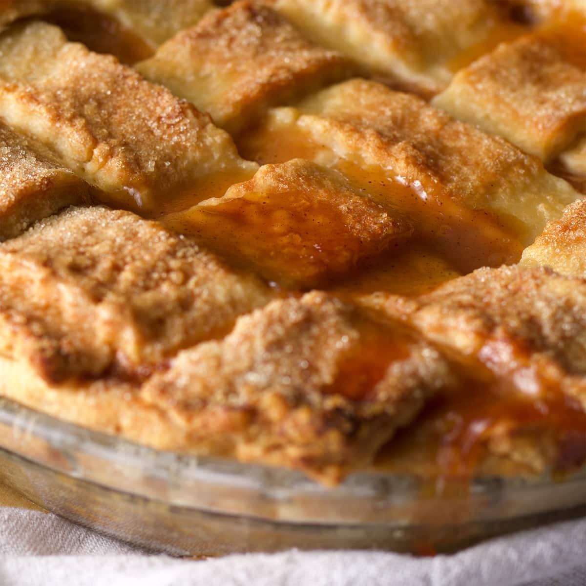 A close up photo of a freshly baked peach pie so you can see the peach pie filling bubbling up in between the gaps in the lattice crust.