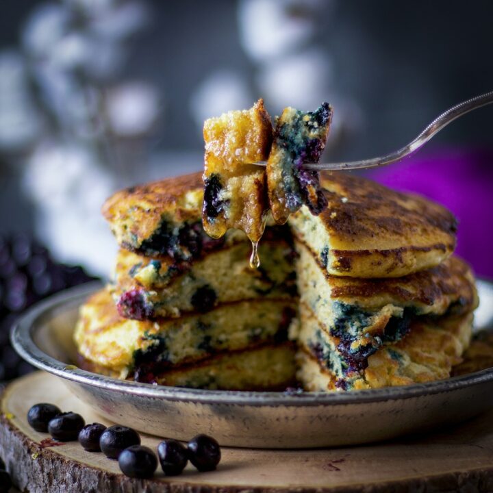 A fork with a bite of fluffy blueberry pancakes on it, syrup dripping from the pancakes, and a stack of blueberry pancakes in the background.