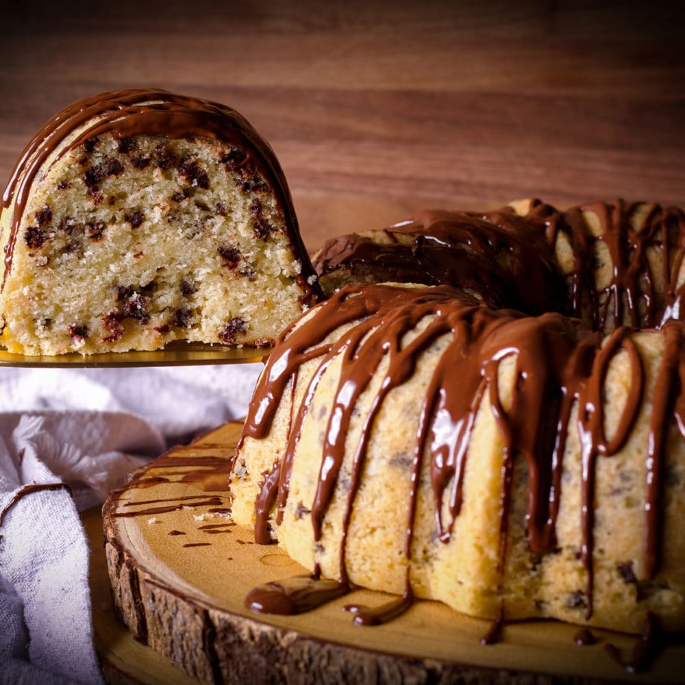 A chocolate chip Bundt Cake on a wood platter with someone using a spatula to lift a slice from the cake.