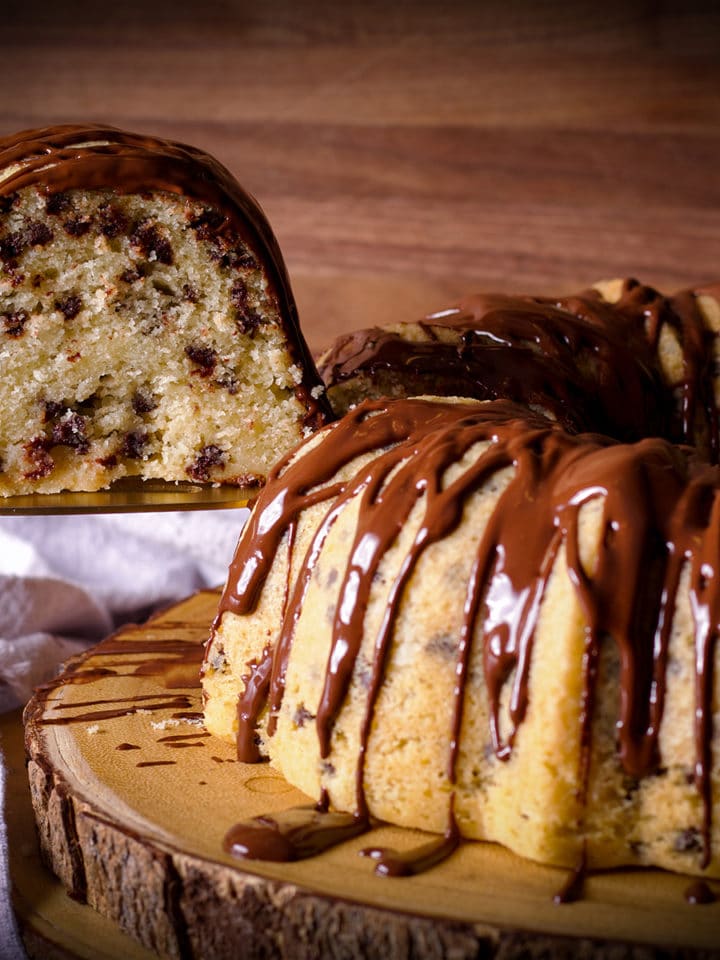 A chocolate chip Bundt Cake on a wood platter with someone using a spatula to lift a slice from the cake.