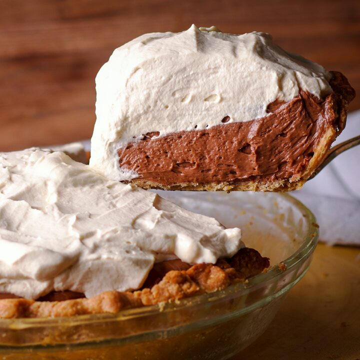 Someone using a spatula to lift a slice of chocolate cream pie from the whole pie so you can see the thick layers of chocolate cream and whipped cream.