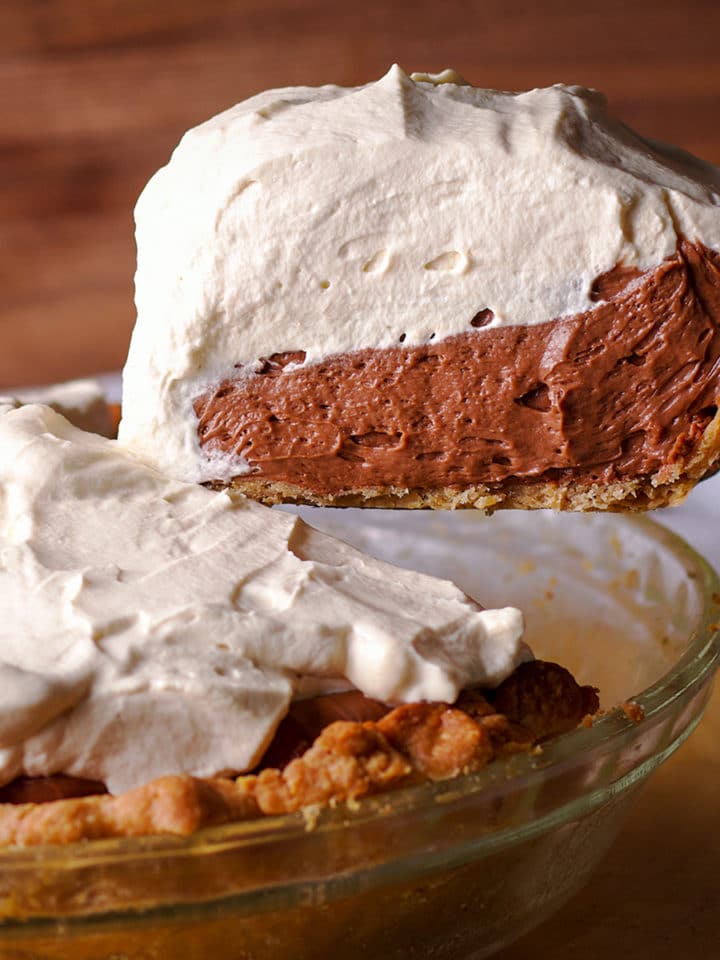 Someone using a spatula to lift a slice of chocolate cream pie from the whole pie so you can see the thick layers of chocolate cream and whipped cream.