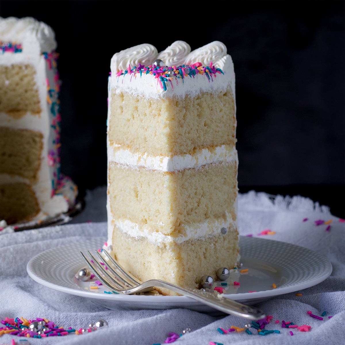 A slice of three layer gluten free vanilla cake frosted with buttercream and decorated with sprinkles on a plate with a fork.