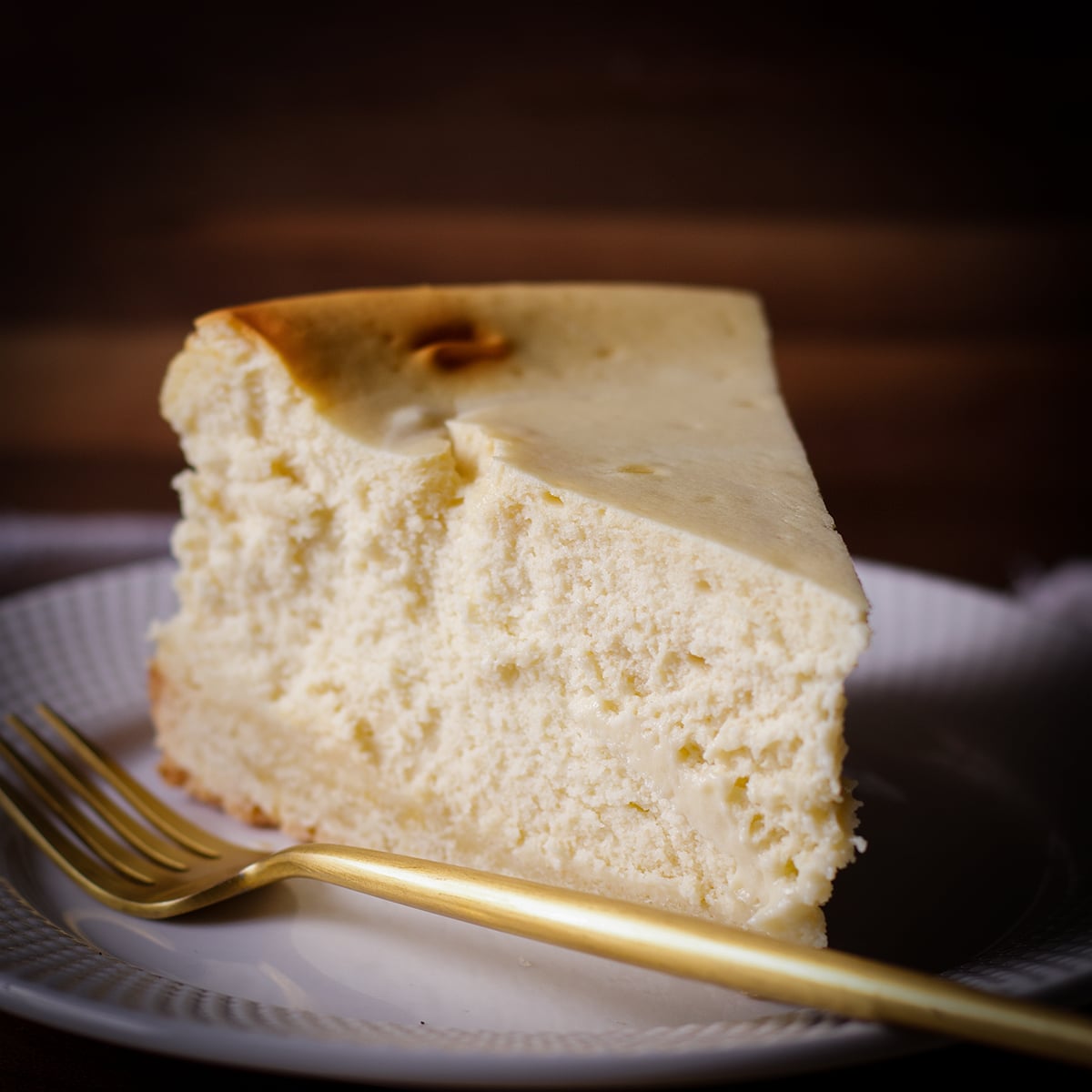 A thick slice of New York Cheesecake on a white plate with a gold fork.