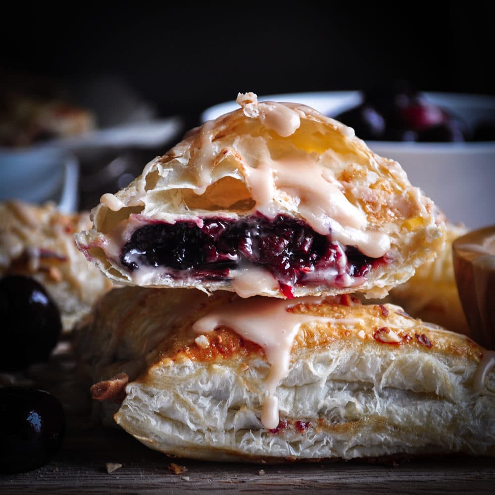 A stack of two cherry turnovers with the top turnover broken in half so you can see the cherry cream cheese filling.
