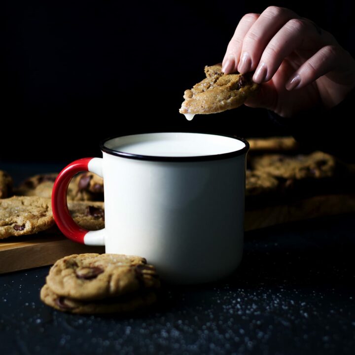 Someone dipping a chocolate chip cookie in a mug that's filled with milk. The mug is surrounded by stacks of chocolate chip cookies.
