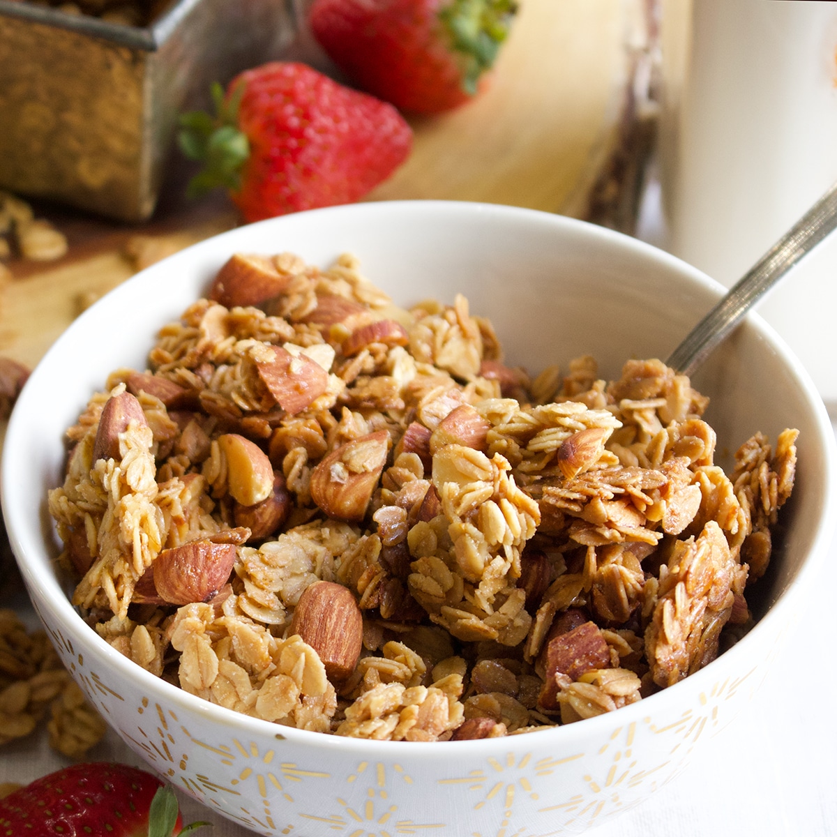 A bowl filled with homemade granola, with milk and fresh strawberries in the background.