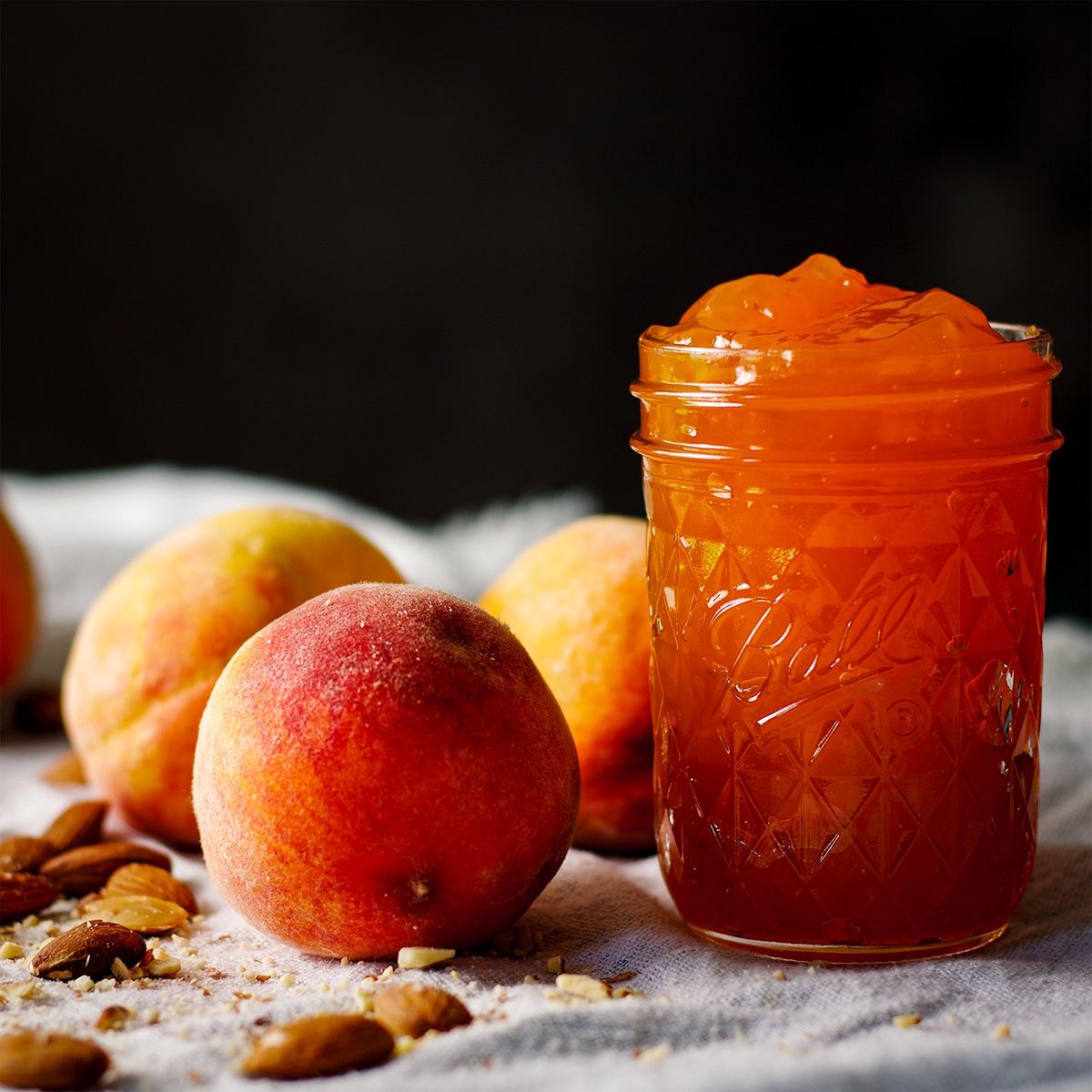 A jar of homemade peach preserves on a table next to fresh peaches and roasted almonds.