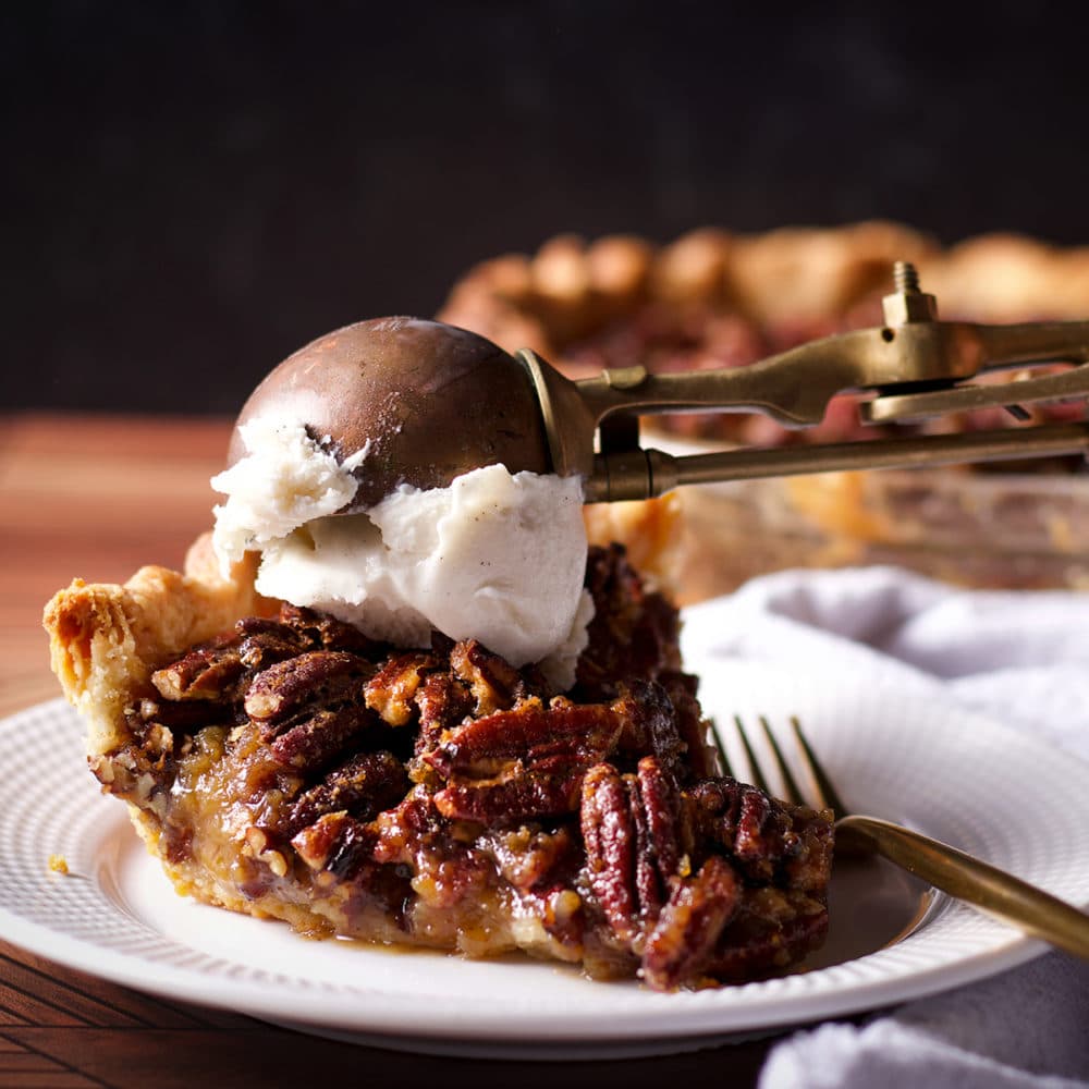 Someone using a old fashioned ice cream scoop to top a slice of pecan pie with a scoop of vanilla ice cream.