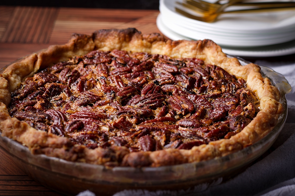 A warm, freshly baked homemade pecan pie resting on a wood table with a stack of plates and forks behind it.