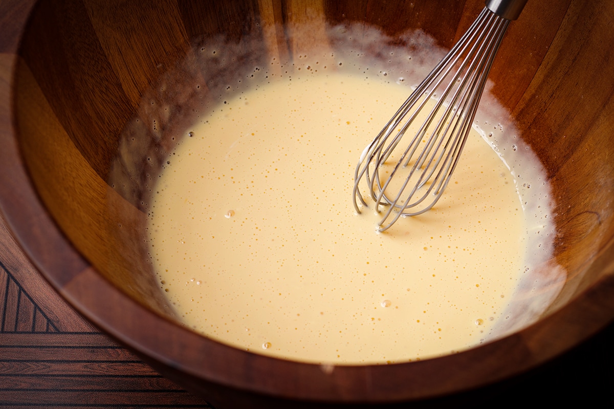 Someone using a wire whisk to blend sour cream into egg yolks.