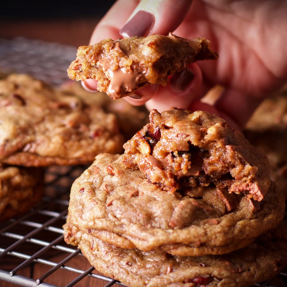 A wire rack with freshly baked chocolate chip pecan cookies cooling on it. One of the cookies has been broken in half so you can see the gooey center.