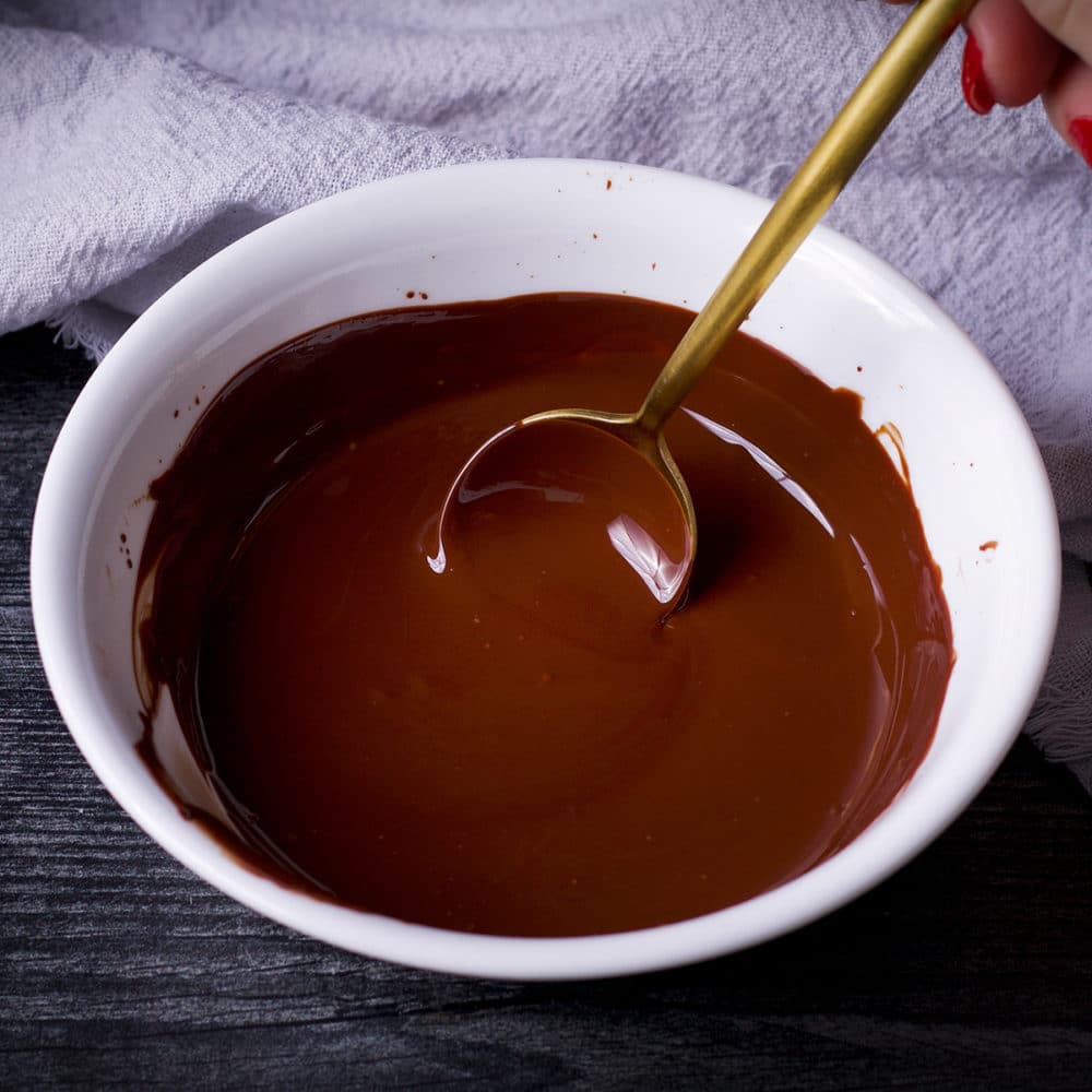 A photo showing someone using a gold spoon to stir melted chocolate in a white bowl.
