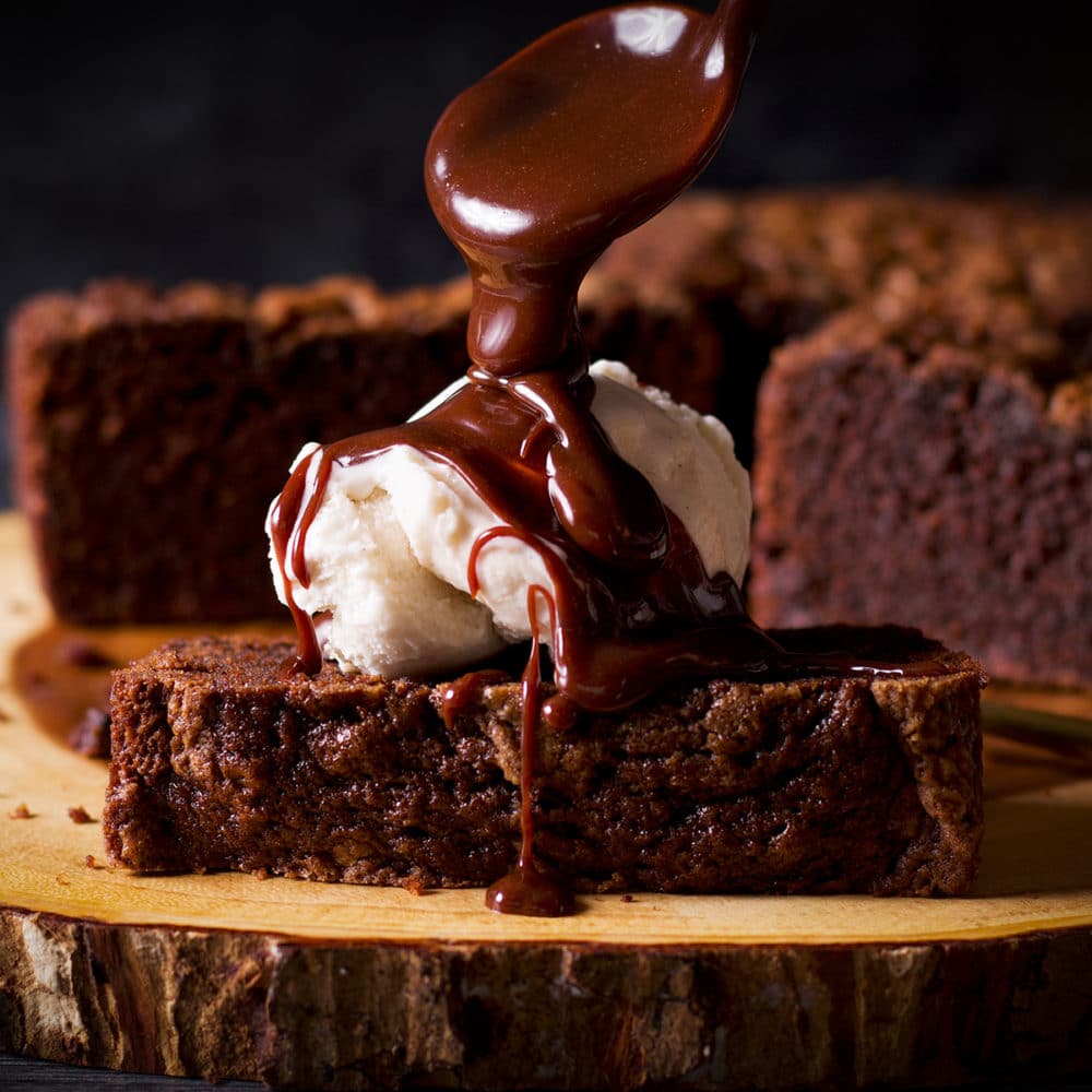 Pouring hot fudge over the top of a piece of chocolate loaf cake topped with vanilla ice cream.