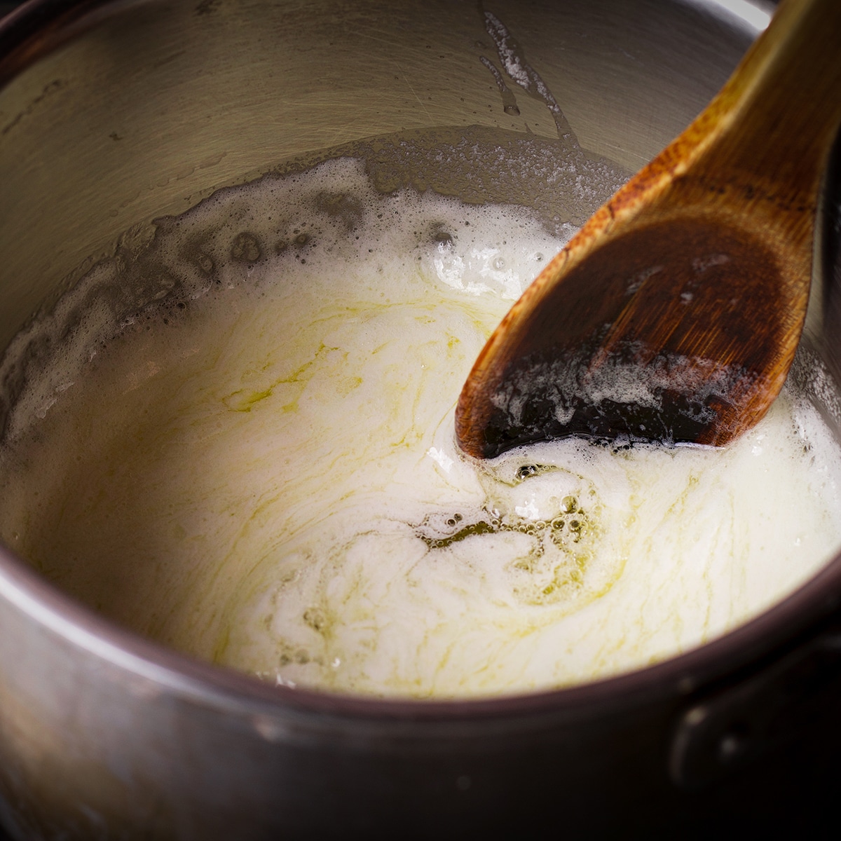 Someone using a wood spoon to stir melted butter in a saucepan. The butter is foamy and just beginning to boil.