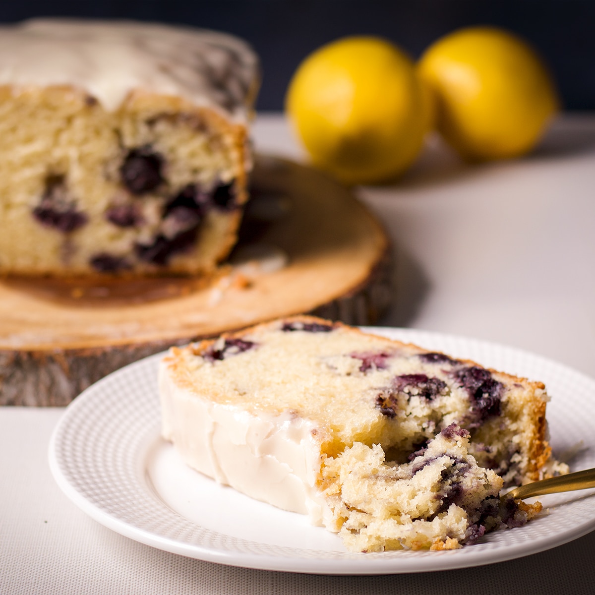 A slice of blueberry loaf cake with lemon icing on a white plate with a gold fork taking a bite of the cake.