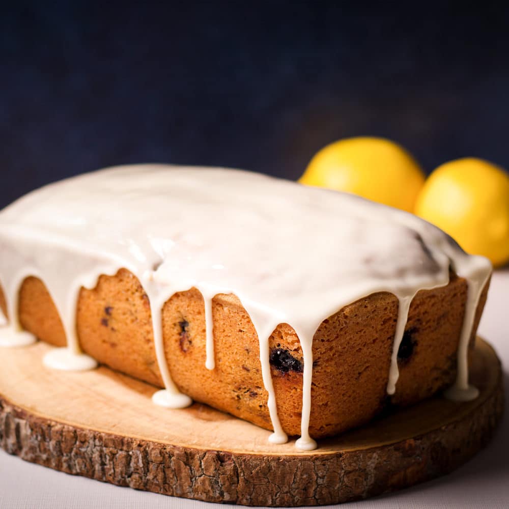 A blueberry loaf cake covered in lemon icing on a wood serving tray.