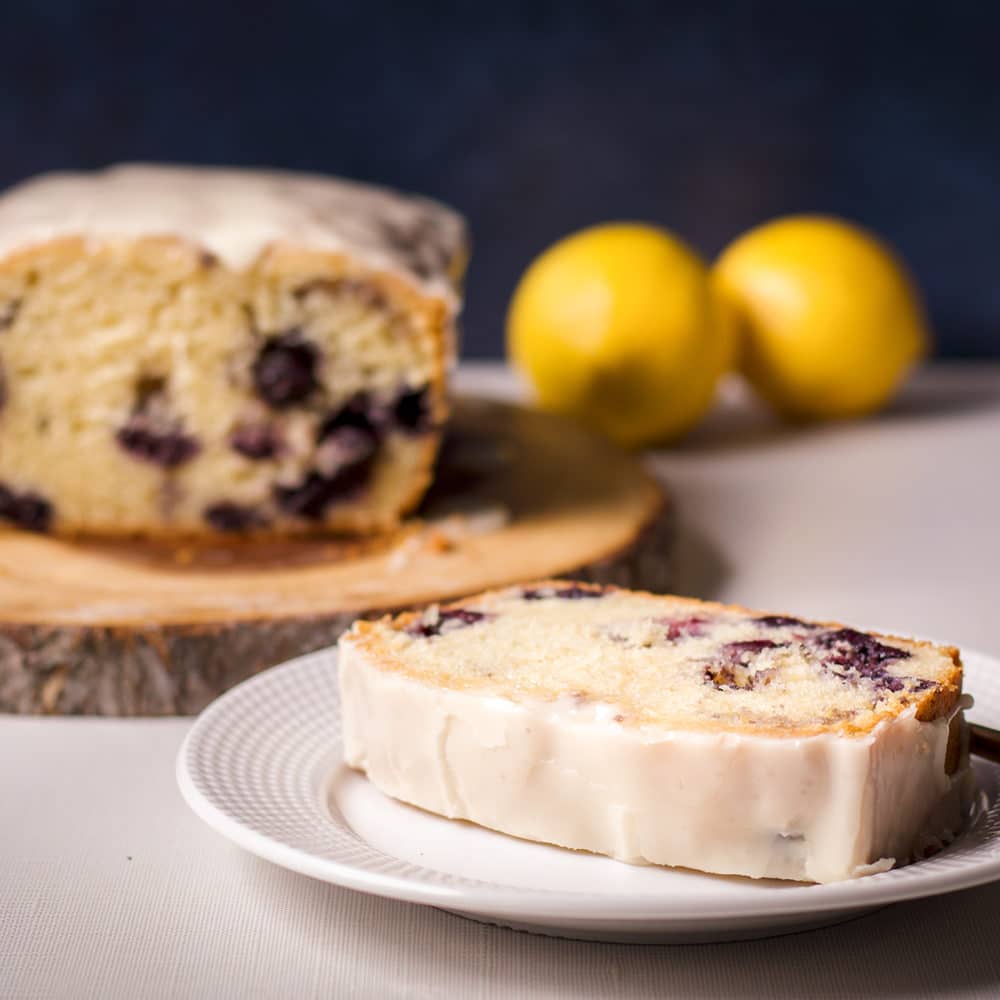 A slice of blueberry loaf cake with lemon icing on a white plate.