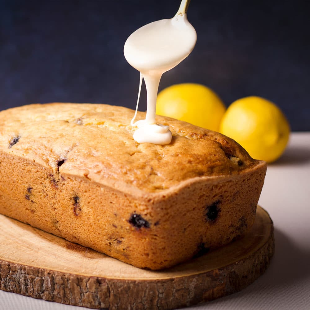 Someone using a spoon to drizzle lemon icing over the top of a blueberry loaf cake.