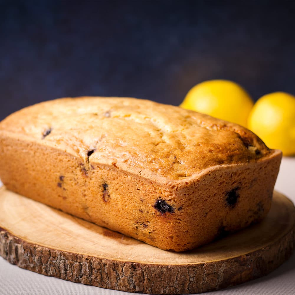 A freshly baked blueberry loaf cake on a wood cutting board with a couple of lemons in the background.
